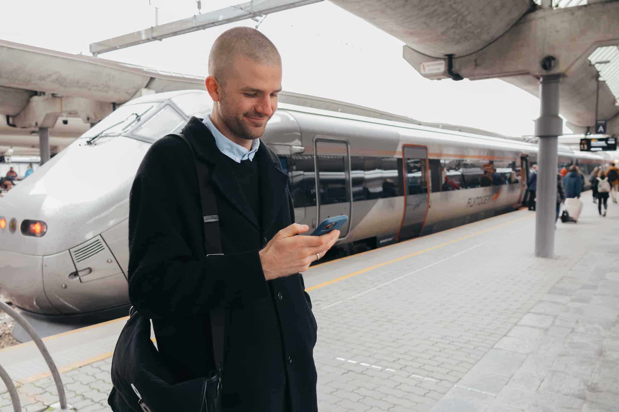 Mann står og ser på telefonen sin med flytoget i bakgrunnen. Foto
